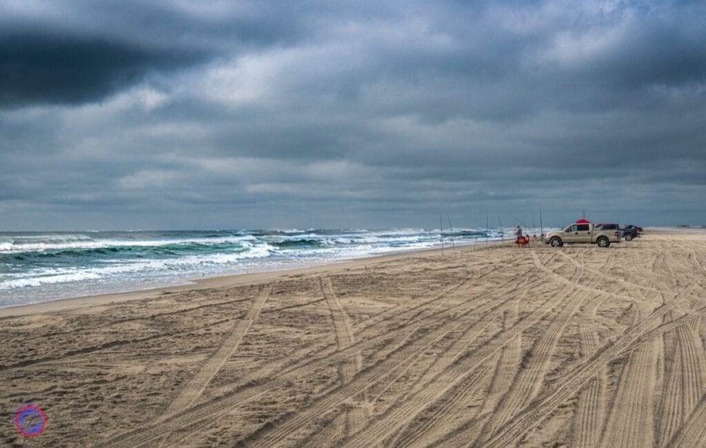 Cloudy beach with trucks and waves, gloomy sky above.