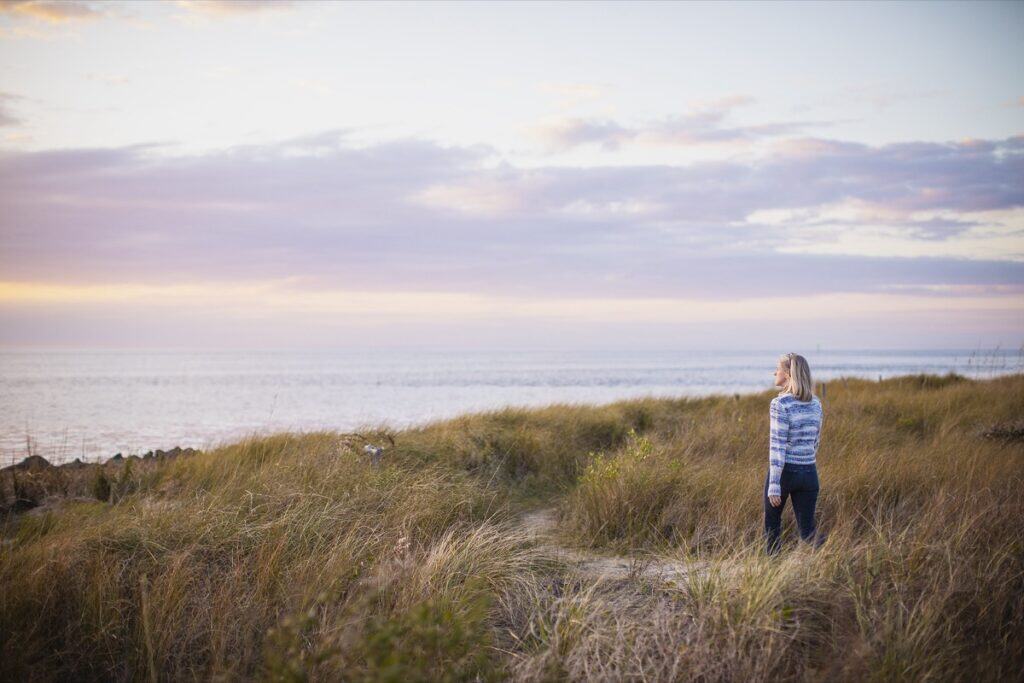 Woman gazing at ocean sunset over grassy dunes.