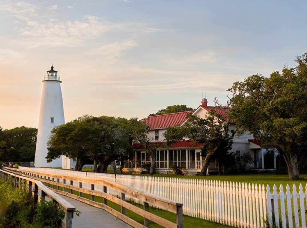 Lighthouse by white fence and house at sunset.