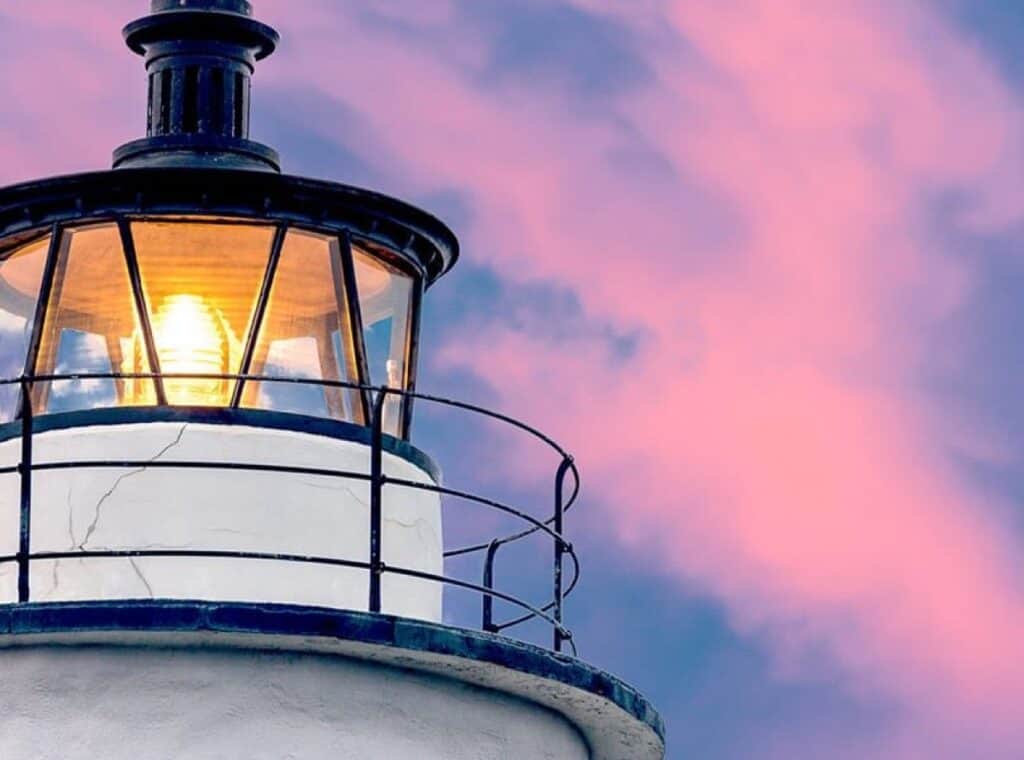 Lighthouse lantern glowing against pink twilight sky.
